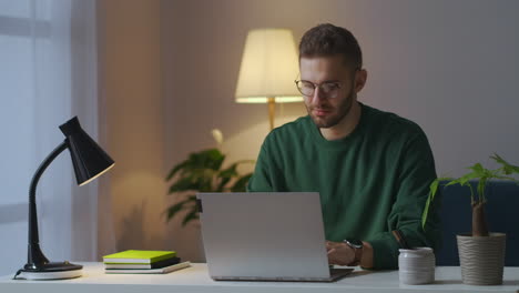 young-man-with-glasses-is-typing-on-laptop-at-evening-at-home-chatting-on-internet-site-medium-portrait-of-intelligent-guy